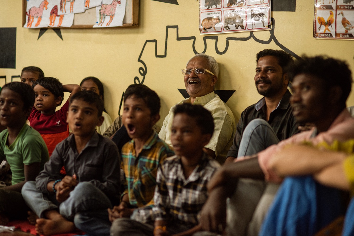 Children and elders laughing together during a learning-centre session, animal charts on the wall